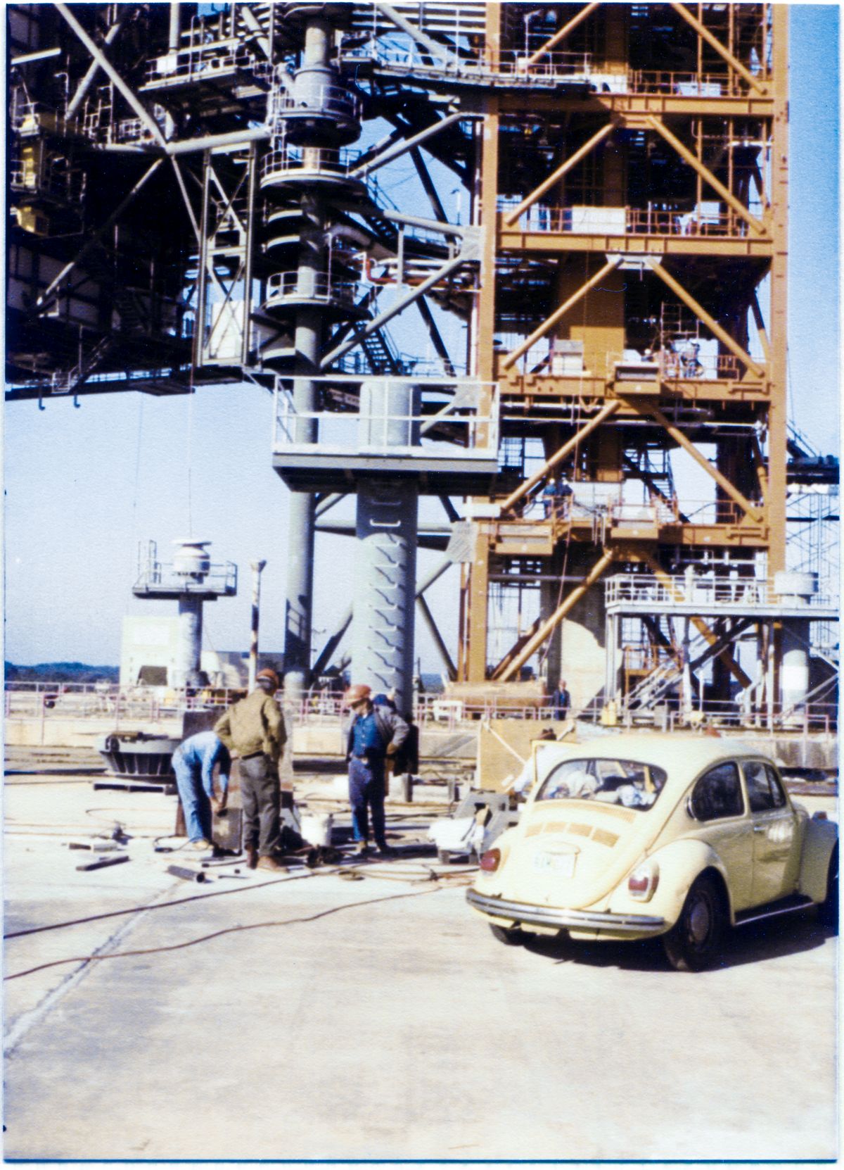 Image 044. With the great dark mass of the Rotating Service Structure at Space Shuttle Launch Complex 39-B at Kennedy Space Center, Florida, looming against the sky in its unmated position to the top left, and the red tones of the Fixed Service Structure extending all the way to the ground to its right, Union Ironworkers from Local 808 working for Wilhoit Steel Erectors prepare a welding setup to put the finishing touch, in the form of a heavy Adjustable Lock Plate, furnished by Sheffield Steel, on the set of Hurricane Tie-downs which will secure the RSS in the mated position against storm winds, if need be. Photo by James MacLaren.